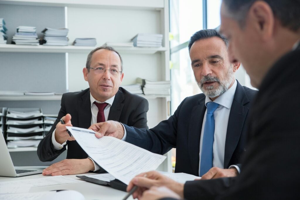 Three businessmen discussing documents in a modern office setting.